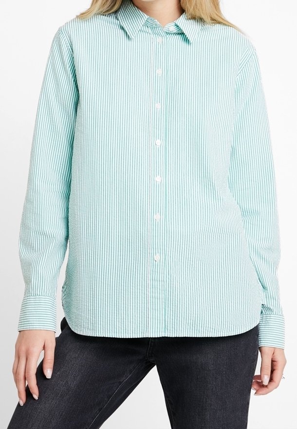Femme portant une chemise boutonnée à rayures verticales vert clair et blanche avec des manches longues et un pantalon sombre, se tenant devant un fond blanc.
