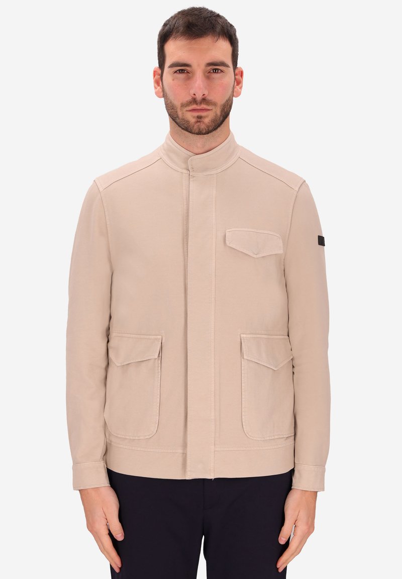 Uomo che indossa una giacca beige con tre tasche a pattina e pantaloni neri, in piedi su uno sfondo bianco.