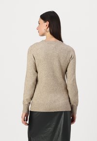 Vaaleanbeige neulepusero, jossa on pitkät hihat, ribbineulospäätös ja pyöreä pääntie, näkyy takaa yksinkertaista valkoista taustaa vasten.