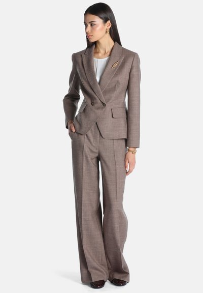 Femme portant un tailleur marron avec pantalon à jambes larges, blouse blanche, broche en or, boucles d’oreilles et bracelet, debout, une main dans la poche, regardant vers la gauche.