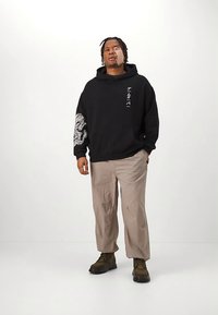 Schwarzer Hoodie mit Fronttasche, versehen mit weißen Drachengrafiken auf dem Ärmel und japanischen Schriftzeichen. Helle beige, locker sitzende Hose und braune Stiefel.