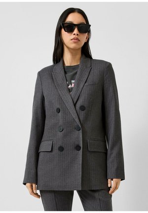 Femme portant un blazer gris foncé à fines rayures, à double boutonnage, surdimensionné, avec des boutons noirs et un pantalon assorti, ainsi que des lunettes de soleil noires.