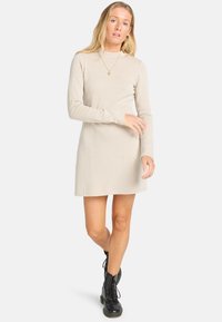 Abito beige a coste con maniche lunghe, corpo aderente, gonna svasata e scollatura alta. Indossato con stivaletti neri con lacci.