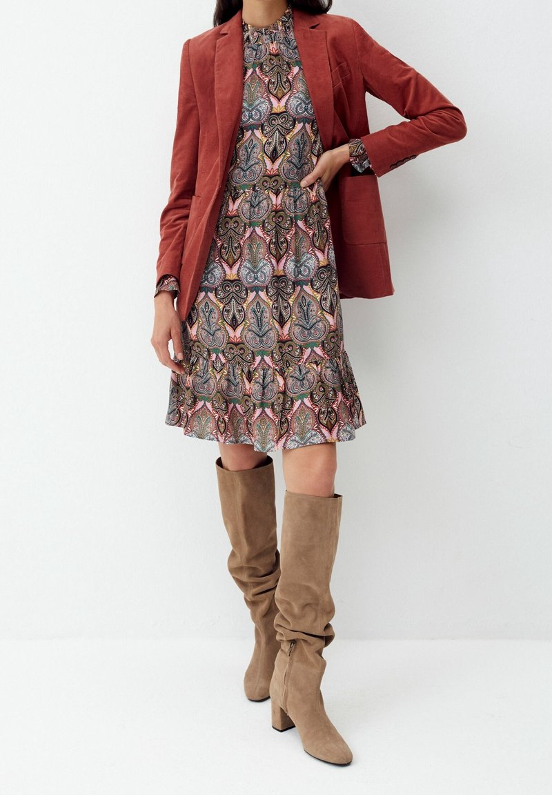 Femme portant une robe à motifs, un blazer couleur rouille et des bottes en daim beige hauteur genoux, debout sur un fond blanc.
