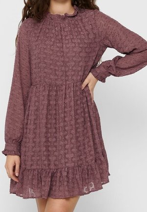 Woman wearing a long-sleeve, mauve textured dress with ruffled cuffs, high neckline, and a ruffled hem, posing with one hand on hip.
