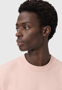 Ροζ ανοιχτόχρωμο φούτερ με στρογγυλή λαιμόκοψη, απαλή υφή και κοντά μανίκια, που φοράει ένα άτομο με κοντά, σγουρά μαλλιά.