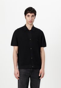 Mlad moški z kratkimi temnimi lasmi, oblečen v črn kratkoslečen srajčni pulover in temno sivo jeans hlače, stoji proti enobarvni beli ozadju.