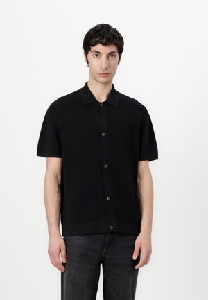 Ung mann med kort mørkt hår som har på seg en svart kortermet skjorte og mørkegrå jeans, står mot en enkel hvit bakgrunn.