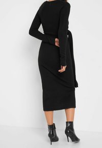 Czarna dzianinowa sukienka z długimi rękawami i związanym detalem w talii, o dopasowanej sylwetce, która sięga poniżej kolan. Błyszczące czarne botki za kostkę.