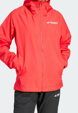 Rote TERREX Regenjacke mit Frontreißverschluss und Kapuze, getragen mit schwarzen TERREX-Hosen, Hände in den Jackentaschen.