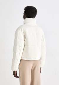 Doudoune en velours côtelé blanc avec un col montant, un design matelassé et une texture côtelée, associée à un pantalon beige, vue de dos.