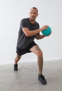 Graues Sport-T-Shirt, schwarze Shorts und schwarze Turnschuhe. Hält einen türkisfarbenen Gymnastikball und hat geometrische Tattoo-Designs an den Beinen.