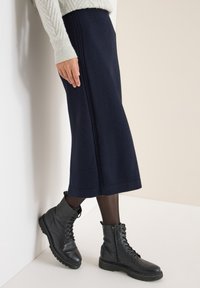 Femme portant une jupe midi bleu marine, des collants noirs transparents, des bottines à lacets noires et un pull en tricot torsadé crème devant un mur clair.