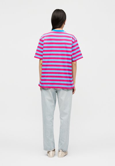 Άτομο με μακριά σκουρόχρωμα μαλλιά που φοράει ροζ και μπλε οριζόντια ριγέ μπλουζάκι, ανοιχτό μπλε τζιν και μπεζ παπούτσια, στέκεται με την πλάτη γυρισμένη.