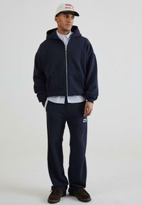 Marineblå zip-up hoodie med kængurulommer, parret med matchende bukser. Outfittet har en stripet skjorte under og brune sko.
