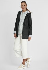 Black rain jacket with a hood, front zipper, and pockets, worn over a blue-striped shirt and white cropped pants, complemented by beige sneakers.