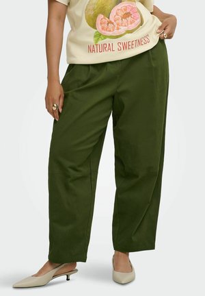 Personne portant un pantalon large vert olive, des talons slingback beiges et un t-shirt clair avec un motif pamplemousse et le texte "Natural Sweetness".