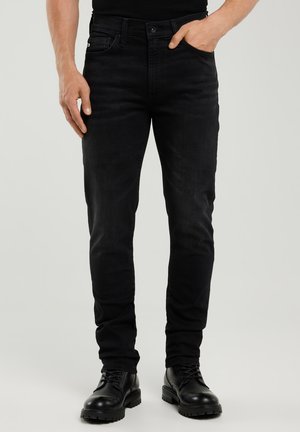 Hombre con vaqueros negros de corte ajustado y botas negras con cordones, con una mano en el bolsillo, frente a un fondo claro y liso.