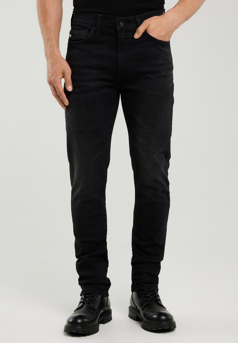 Bărbat purtând blugi negri slim-fit cu bocanci negri cu șireturi, o mână în buzunar, pe un fundal simplu și deschis la culoare.