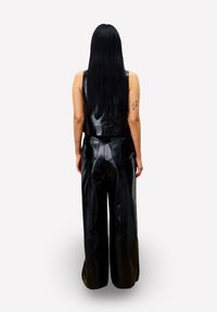 Person mit langen schwarzen Haaren, der ein ärmelloses schwarzes Lederoberteil und weite schwarze Lederhosen trägt, steht mit dem Rücken zur Kamera vor einem weißen Hintergrund.