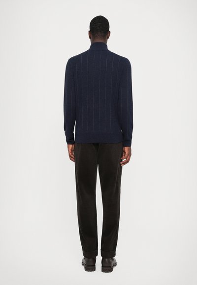 Granatowy sweter z pionowymi paskami, wysoki kołnierz i długie rękawy, w połączeniu z ciemnymi spodniami sztruksowymi i brązowymi botkami. Minimalistyczny design.