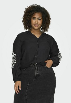 Chemise noire à boutons avec manches longues, ornée de motifs décoratifs blancs sur les manches, assortie d'une jupe en denim noir.