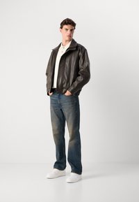 Chaqueta bomber de cuero marrón con cremallera frontal, llevada sobre una camisa blanca con cuello, combinada con unos vaqueros azules desgastados y zapatillas blancas.