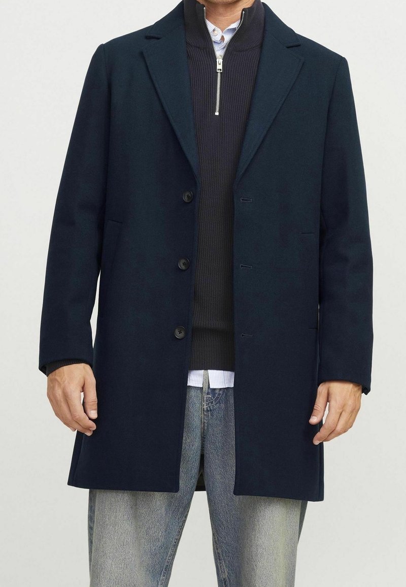 Marinblå ullrock med slagkrage, fyra knappar och sidofickor, lager på lager över en stickad tröja och skjorta, tillsammans med ljusblå jeans.