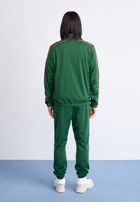 Survêtement vert en tissu lisse, avec un col zippé et des rayures dorées sur les manches. Assorti à des baskets blanches.