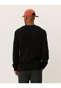 Czarny sweter o strukturalnej fakturze z prążkowanymi mankietami i dołem, z okrągłym dekoldem. Modelka ma na sobie kapelusz w kolorze rdzy.