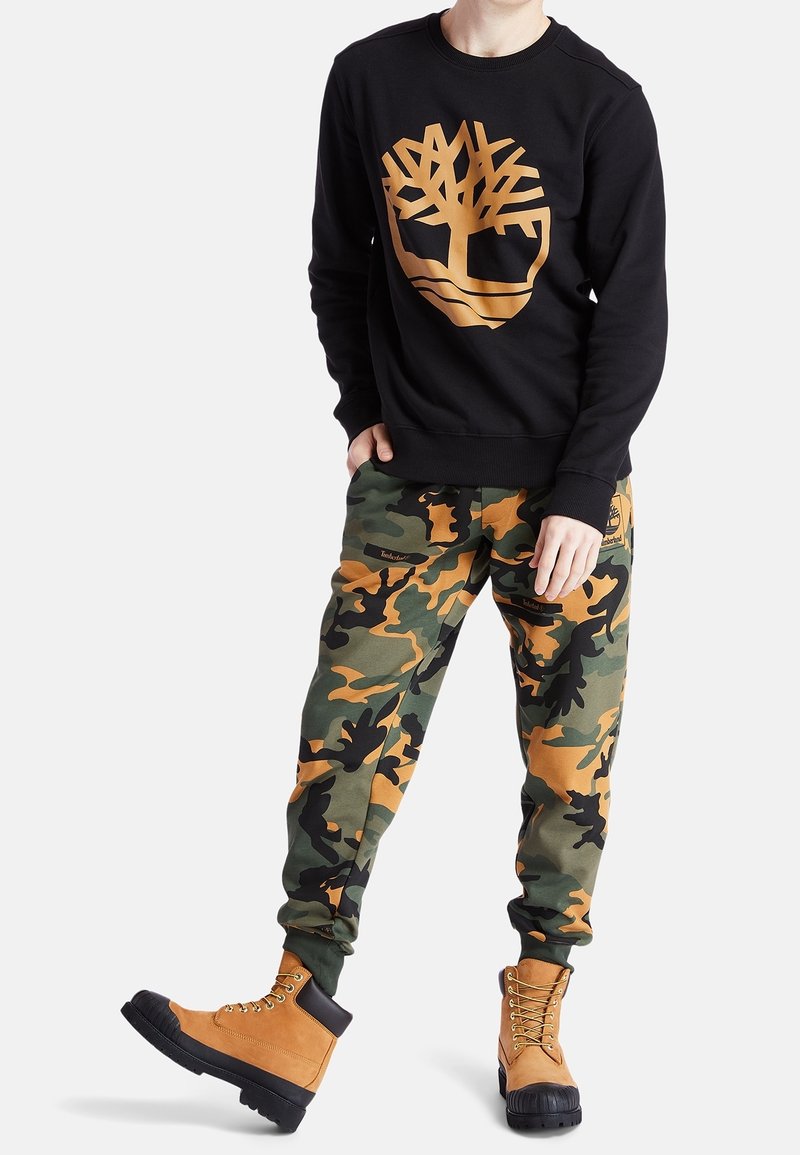 Persona vestida con sudadera negra con logo de árbol en bronce, pantalones de camuflaje y botas tan con cordones, de pie con un pie ligeramente elevado.