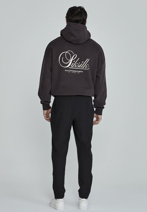 Sweat à capuche noir avec un grand texte graphique blanc au dos, associé à un pantalon noir et des baskets blanches, affichant une coupe décontractée et un style casual.