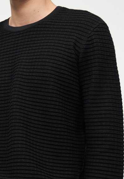 Άνδρας που φοράει μαύρο μακρυμάνικο πουκάμισο με υφή και οριζόντιο ριμπ σχέδιο, εμφανιζόμενος από τον λαιμό έως τη μέση, μπροστά σε απλό φόντο.