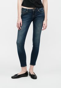 Mujer vestida con jeans ajustados azul oscuro, top corto negro con botones y bailarinas negras, de pie sobre un fondo blanco liso.