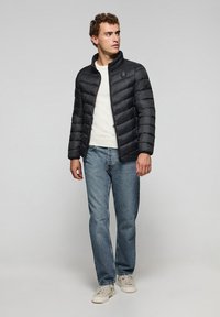 Sort pufferjakke med stående krave og chevron quiltet mønster, båret over en lys sweater, parret med blå jeans og hvide sneakers.