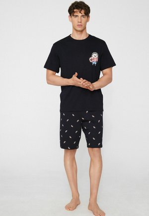 Hombre de pie descalzo vistiendo una camiseta negra de manga corta con un pequeño gráfico de erizo y pantalones cortos negros a juego con un patrón repetido de erizos.
