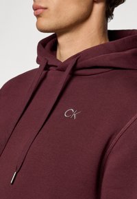 Burgundi színű kapucnis pulóver, állítható kapucnival, amelyen egy kis hímzett "CK" logó található. Az anyag lágy és sima tapintású.