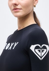 Κοντινό πλάνο γυναίκας που φοράει μακρυμάνικο μαύρο τοπ με λευκή επιγραφή «Roxy» στο στήθος και λογότυπο σε σχήμα καρδιάς στο μανίκι.