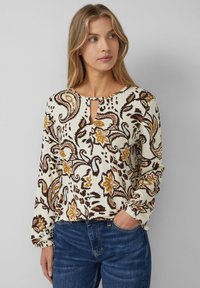 Kobieta z blond włosami w kremowej bluzce z brązowym i musztardowym wzorem kwiatowym w stylu paisley oraz niebieskich dżinsach, jedna ręka w kieszeni, patrzy w prawo.