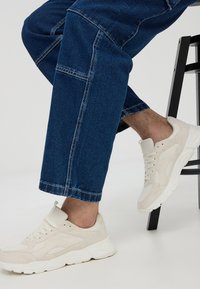 Beżowe sportowe trampki z górą wykonaną z teksturowanej tkaniny, lekkim amortyzowaniem i białą gumową podeszwą. Noszone z ciemnoniebieskimi jeansami o prostych nogawkach.