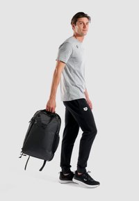 Sort rygsæk med lynlåse, bærestrop og logo; grå t-shirt; sorte joggers; sorte og grå træningssko; neutral baggrund.