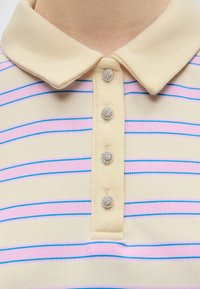 Camicia beige con colletto con righe orizzontali rosa e blu, con quattro bottoni rotondi scintillanti sulla patta frontale.