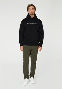 Sudadera negra con bolsillo delantero y capucha con cordón, que presenta el logo "SAIL RACING" en blanco. Combinada con pantalones verde oliva y zapatillas blancas.