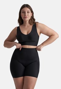 Donna che indossa un reggiseno sportivo nero senza cuciture e pantaloni a vita alta modellanti, mentre regola l'elastico contro uno sfondo bianco semplice.