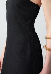 Schwarzes Leinenkleid mit tailliertem Oberteil und gerafften Details an der Seite. Verfügt über einen dezenter Reißverschluss und eine glatte, strukturierte Oberfläche.