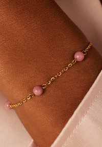 Gouden armband met drie roze kralen, tegen een textuur van huid achtergronden. Gladde kralen contrasteren met de schakels van de ketting.