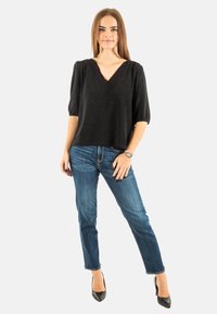 Top corto nero con maniche a sbuffo e scollatura a V in un tessuto lavorato, abbinato a jeans blu aderenti e tacchi neri a punta. Accessori minimi.