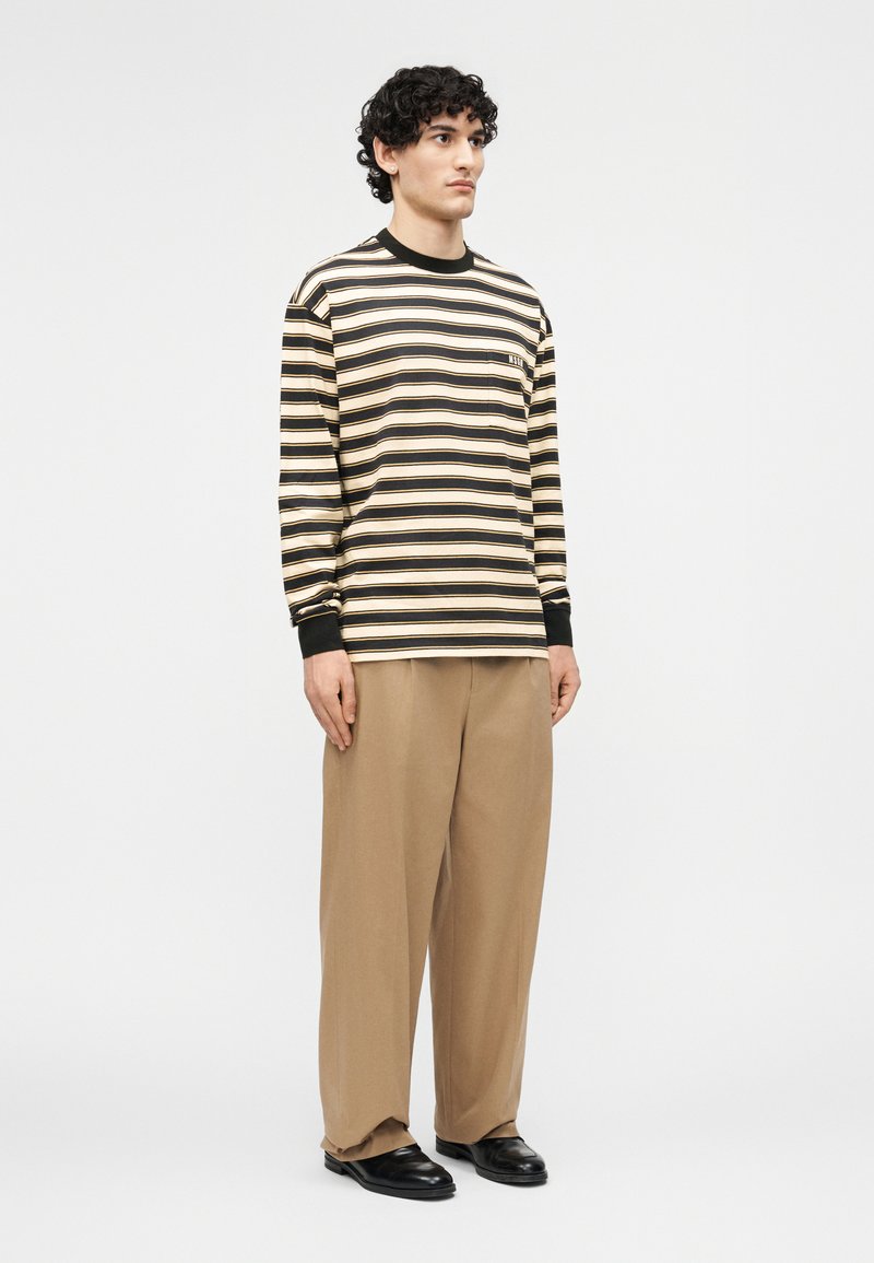 Man met krullend haar, gekleed in een zwart-beige gestreept shirt met lange mouwen, losse beige broek en zwarte schoenen, staand tegen een effen achtergrond.
