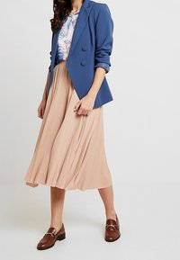 Blazer blu doppiopetto sopra una camicetta floreale, abbinato a una gonna plissettata rosa e mocassini marroni. Nota: la gonna è di lunghezza media e fluida.