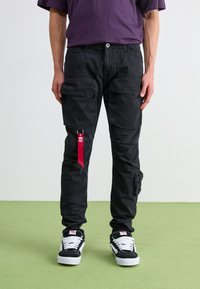 Black cargo pants with multiple pockets, a fabric loop with a red accent, and a relaxed fit, paired with black and white sneakers.